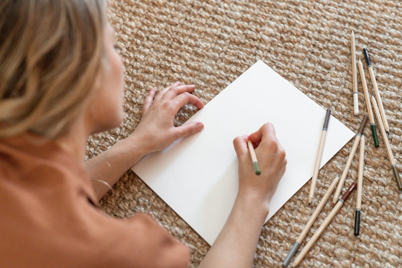 Top view of a woman sketching on paper with colored pencils on a textured mat.